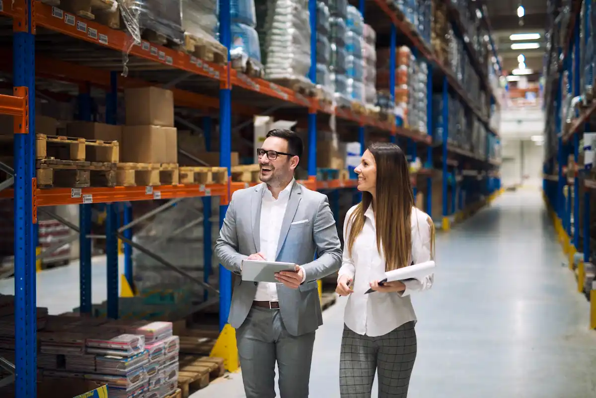 Logistics managers discussing material supply inventory tracking inside a large industrial warehouse.